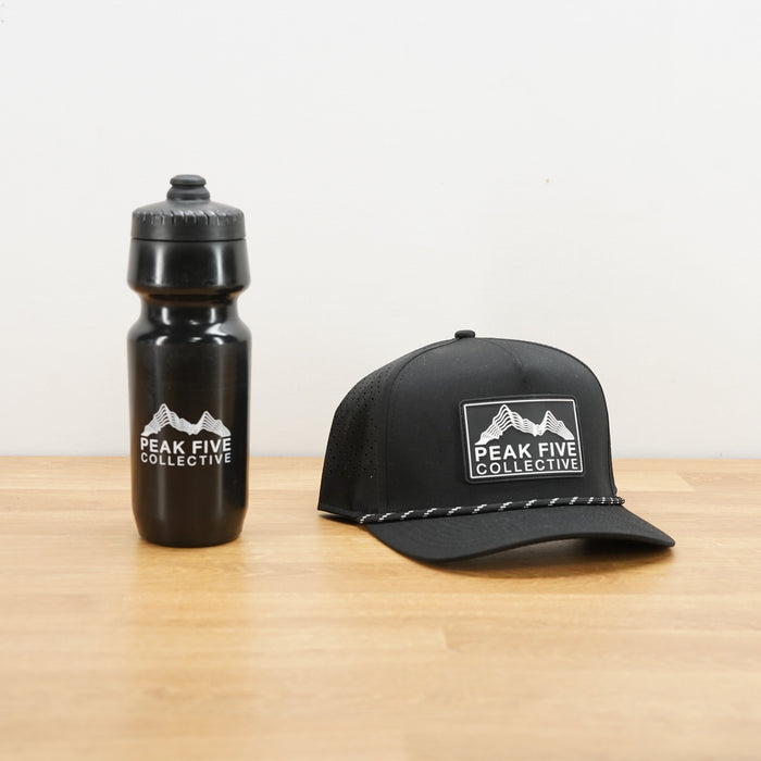 A black water bottle and a black baseball cap, both printed with the Peak Five Collective logo, displayed on a light wood tabletop against a white wall.