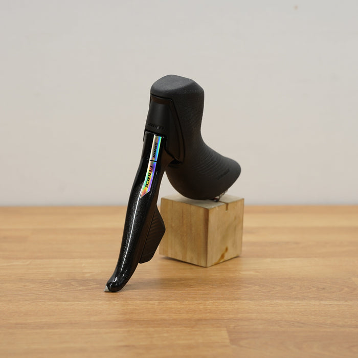 A black SRAM shift lever displayed on a small wooden block on a light wood tabletop against a plain light gray wall.