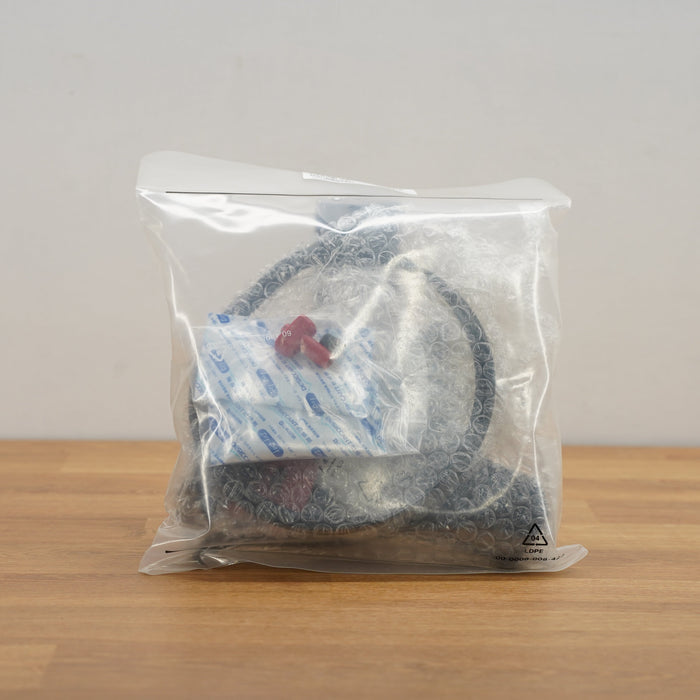 A clear plastic bag containing a black coiled cable or hose with a small red-and-white tag, placed on a light wood surface against a plain light gray wall.
