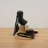 A black SRAM road bike shifter lever mounted on a small wooden block, with its black cable coiled in front on a light wood tabletop against a plain off-white wall.