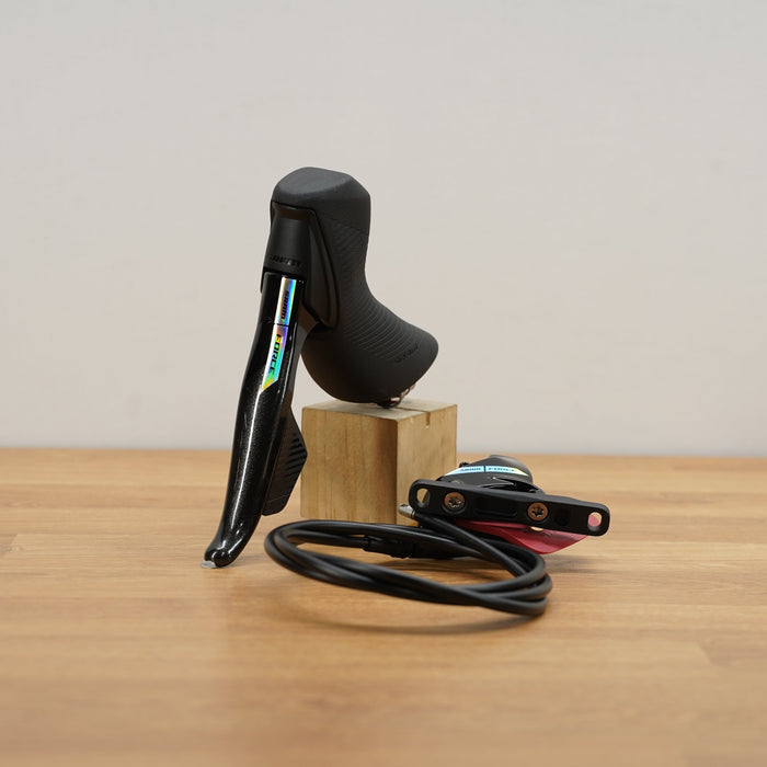 A black SRAM road bike shifter lever mounted on a small wooden block, with its black cable coiled in front on a light wood tabletop against a plain off-white wall.