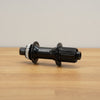 A Shimano black bicycle hub with a silver freehub body, resting on a light brown wooden surface against a neutral beige background.