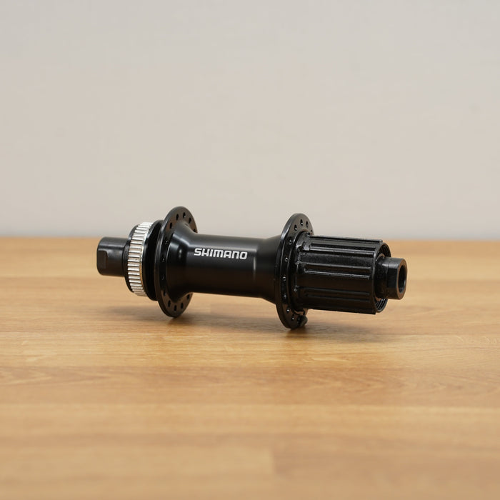 A Shimano black bicycle hub with a silver freehub body, resting on a light brown wooden surface against a neutral beige background.