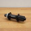 A Shimano black bicycle hub with a silver splined end, resting on a light brown wooden surface against a neutral gray background.