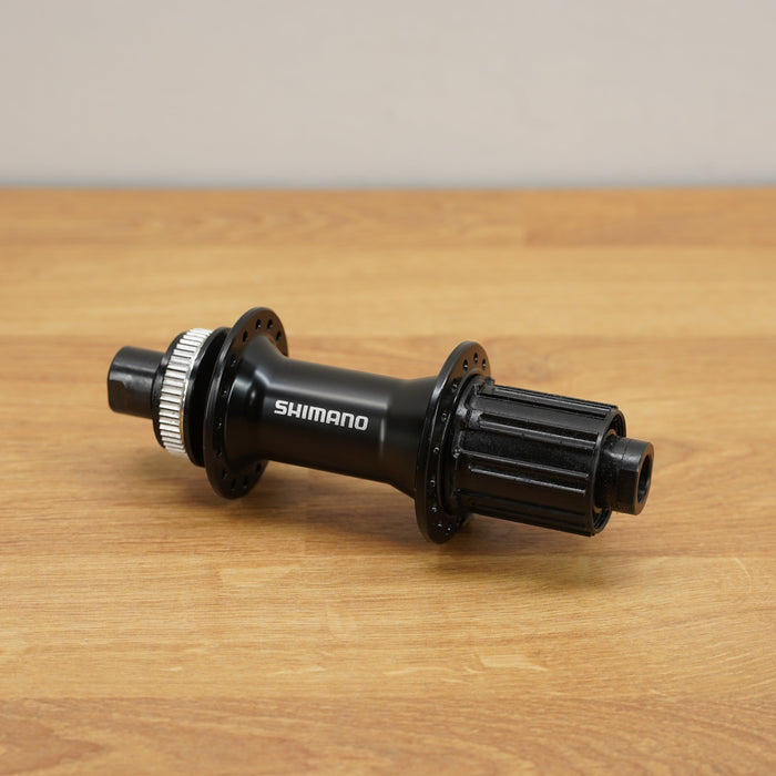 A Shimano black bicycle hub with a silver splined end, resting on a light brown wooden surface against a neutral gray background.