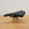 A black bicycle saddle displayed on a small wooden block stand over a light wood tabletop against a plain off-white wall.