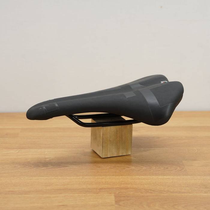 A black bicycle saddle displayed on a small wooden block stand over a light wood tabletop against a plain off-white wall.