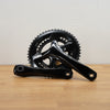 A black bicycle crankset, displayed on a light wood surface against a plain off-white wall.