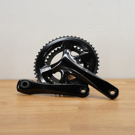 A black bicycle crankset, displayed on a light wood surface against a plain off-white wall.