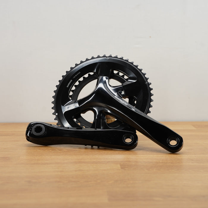 A black bicycle crankset, displayed on a light wood surface against a plain off-white wall.