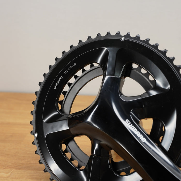 A close-up of a black Shimano bicycle crank arm and chainring assembly resting on a wooden surface against a neutral beige background.