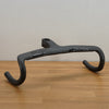 A dark gray carbon fiber road bike handlebar, displayed on a light wood tabletop against a plain off-white wall.