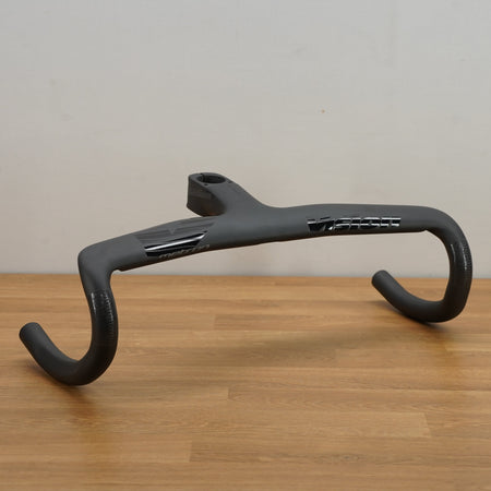 A dark gray carbon fiber road bike handlebar, displayed on a light wood tabletop against a plain off-white wall.