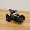 A bicycle rear derailleur in matte black and dark gray metal, photographed on a light wood tabletop against a plain beige wall.
