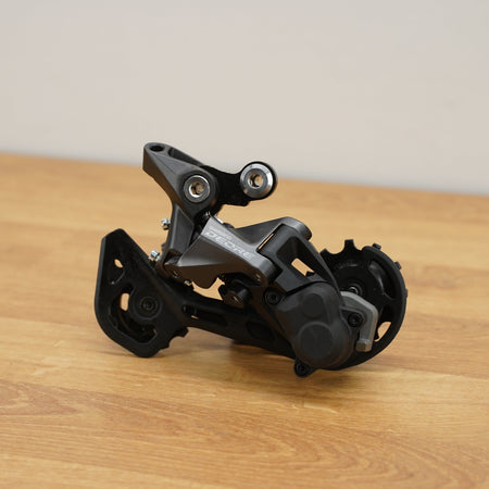 A bicycle rear derailleur in matte black and dark gray metal, photographed on a light wood tabletop against a plain beige wall.