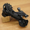 A black bicycle rear derailleur mounted on a small wooden block, photographed on a light wood tabletop.