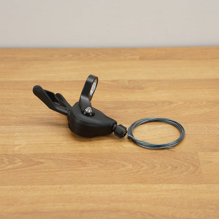 A black Shimano bicycle shift lever with an attached cable resting on a light brown wooden floor against a neutral beige wall.