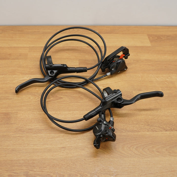 A pair of black bicycle brake levers with attached brake cables, laid out on a light brown wooden floor against a white wall.