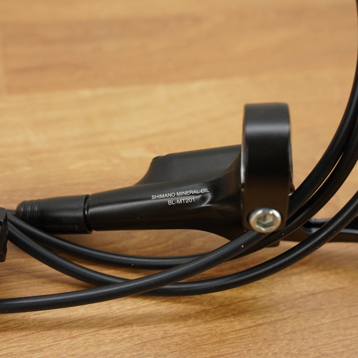 A close-up of a black Shimano hydraulic disc brake lever with its attached cable, resting on a light brown wooden surface.