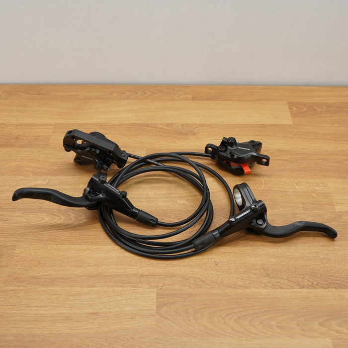 Black bicycle brake lever set with a coiled cable and brake caliper, placed on a light wood floor against a neutral wall.