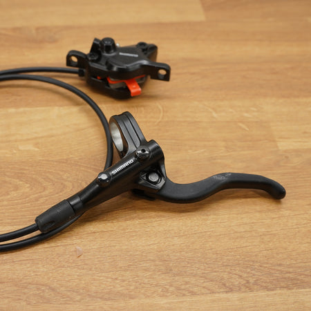 A black Shimano bicycle brake lever with its attached black cable and a matching black brake mount, displayed on a light brown wooden surface.