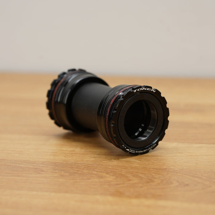 A black bicycle bottom bracket resting on a light brown wooden tabletop against a neutral beige background.