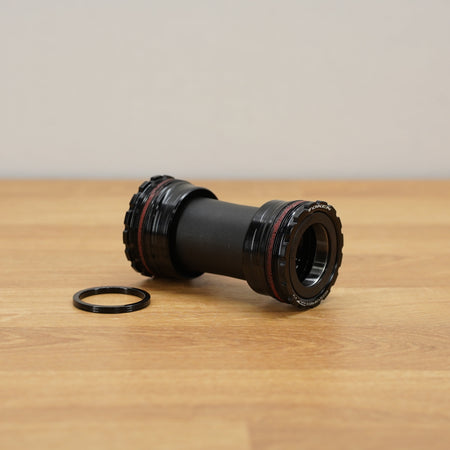 A black bicycle bottom bracket with a small black ring placed beside it on a light wood tabletop against a neutral beige wall.