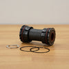 A black bicycle bottom bracket with red accents, shown on a light wood tabletop with two black gaskets and a silver clip in front of it.