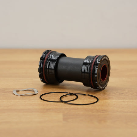 A black bicycle bottom bracket with red accents, shown on a light wood tabletop with two black gaskets and a silver clip in front of it.