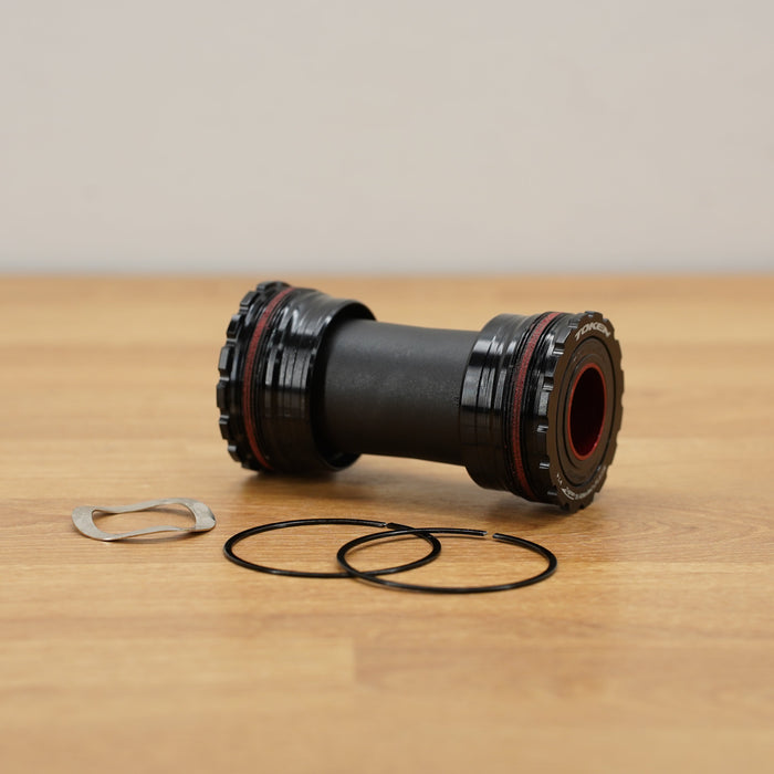 A black bicycle bottom bracket with red accents, shown on a light wood tabletop with two black gaskets and a silver clip in front of it.