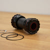 A black bicycle bottom bracket with red accents and white branding, shown on a wooden tabletop with two black seals and a small silver metal piece nearby.