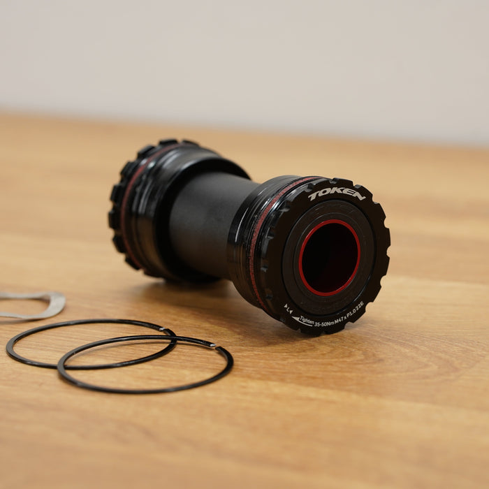A black bicycle bottom bracket with red accents and white branding, shown on a wooden tabletop with two black seals and a small silver metal piece nearby.