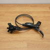 A black Shimano hydraulic disc brake lever assembly with a coiled black cable, resting on a light brown wooden tabletop.