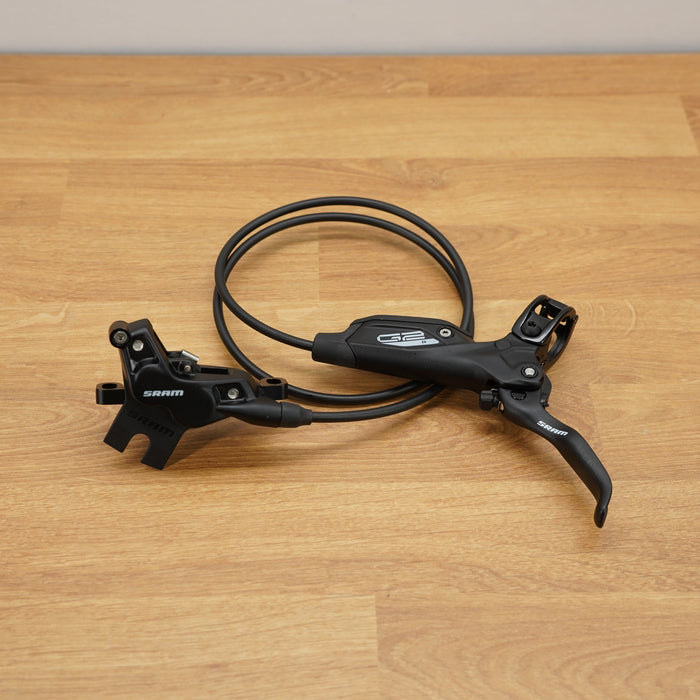 A black Shimano hydraulic disc brake lever assembly with a coiled black cable, resting on a light brown wooden tabletop.