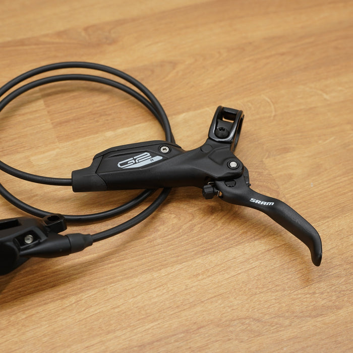 A Shimano SRAM bicycle brake lever and matching brake caliper with a coiled black cable, displayed on a light brown wooden surface.