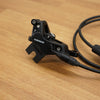 A close-up of a black SRAM hydraulic disc brake caliper with its attached black cable running across a light brown wooden surface.