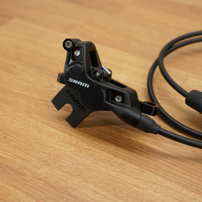 A close-up of a black SRAM hydraulic disc brake caliper with its attached black cable running across a light brown wooden surface.