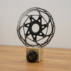 A black metallic disc brake rotor displayed upright on a small wooden block with a black cylindrical base, set on a wooden tabletop against a plain light wall.