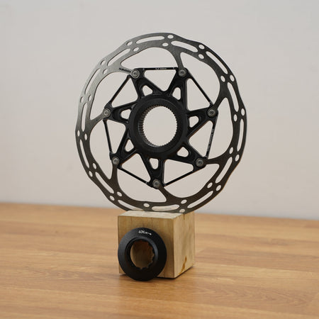A black metallic disc brake rotor displayed upright on a small wooden block with a black cylindrical base, set on a wooden tabletop against a plain light wall.
