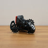 A black and gray bicycle derailleur with a small red accent, resting on a light brown wooden surface against a neutral beige background.