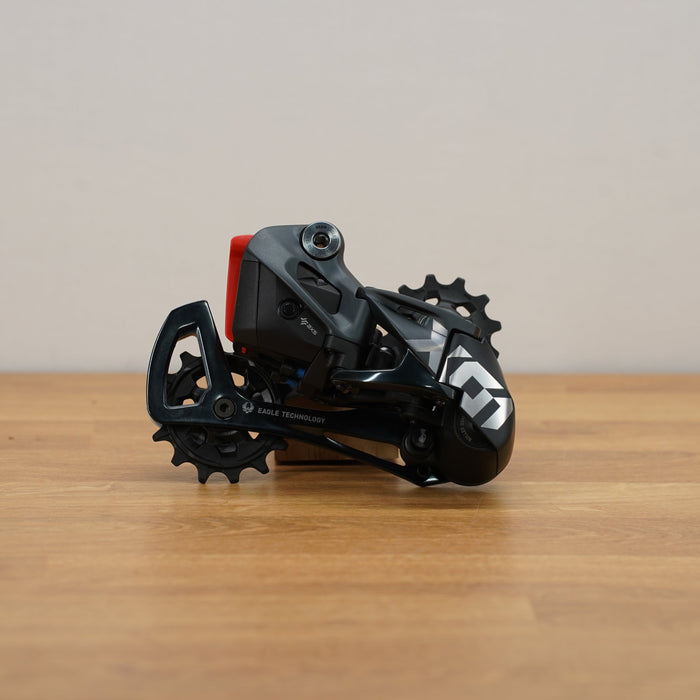 A black and gray bicycle derailleur with a small red accent, resting on a light brown wooden surface against a neutral beige background.