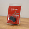 SRAM bicycle accessory in clear blister packaging, featuring a red card insert with the SRAM logo and a small black rectangular component inside, photographed on a wooden surface against a light wall.