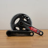 A grey SRAM crankset with a red sticker, resting on a light wood floor against a plain off-white wall.