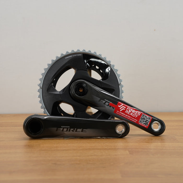 A grey SRAM crankset with a red sticker, resting on a light wood floor against a plain off-white wall.