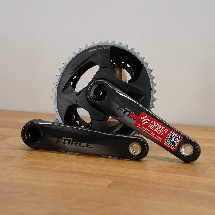 A grey SRAM crankset with a red sticker, resting on a light wood floor against a plain off-white wall.