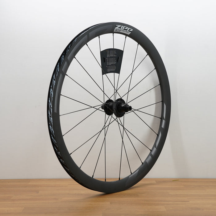 A carbon fiber ZIPP bicycle wheel with a black deep-section rim, thin black spokes, and a black hub, standing upright on a light wood floor against a plain white wall.
