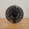 A black multi-sprocket bicycle cassette displayed upright on a light wood surface against a plain off-white wall.