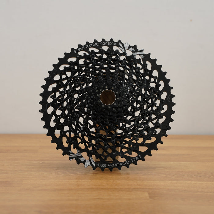 A black multi-sprocket bicycle cassette displayed upright on a light wood surface against a plain off-white wall.