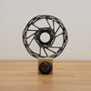 A black metal disc brake rotor displayed upright on a small wooden block stand against a plain light gray wall and wood tabletop.