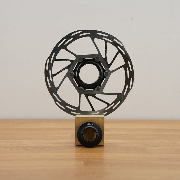 A black metal disc brake rotor displayed upright on a small wooden block stand against a plain light gray wall and wood tabletop.